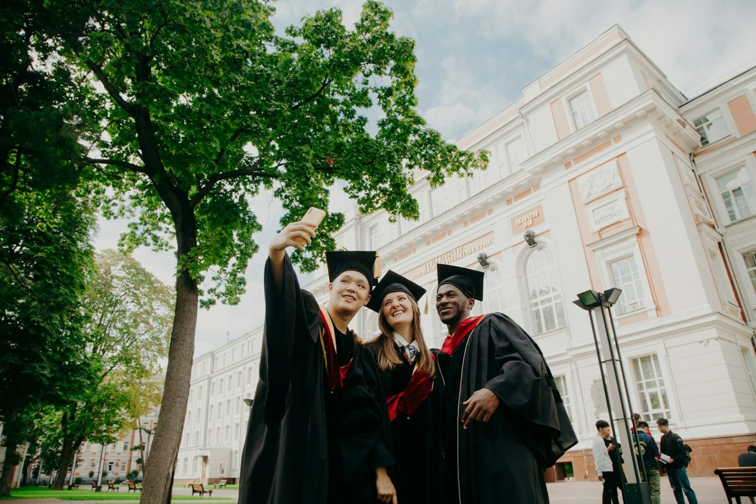 Three graduates in caps and gowns, including an international student, take a selfie in front of a large, ornate building on a sunny day.