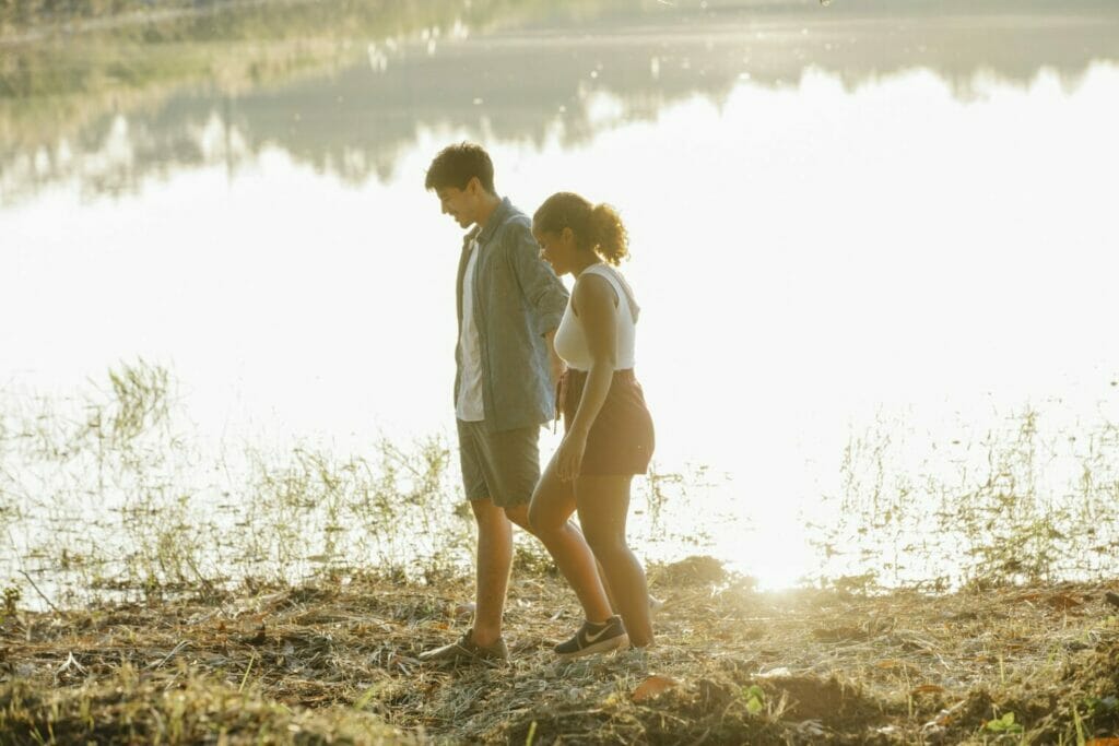 Content young multiethnic couple holding hands while waling on lake shore at sunset