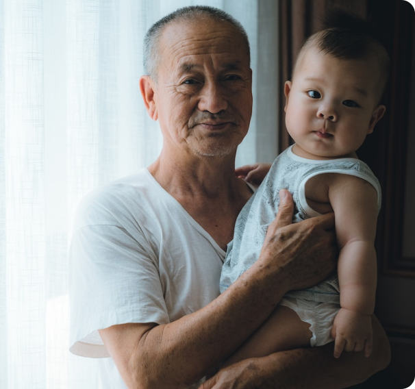 An elderly man holding a baby, both looking toward the camera, near a window with sheer curtains.