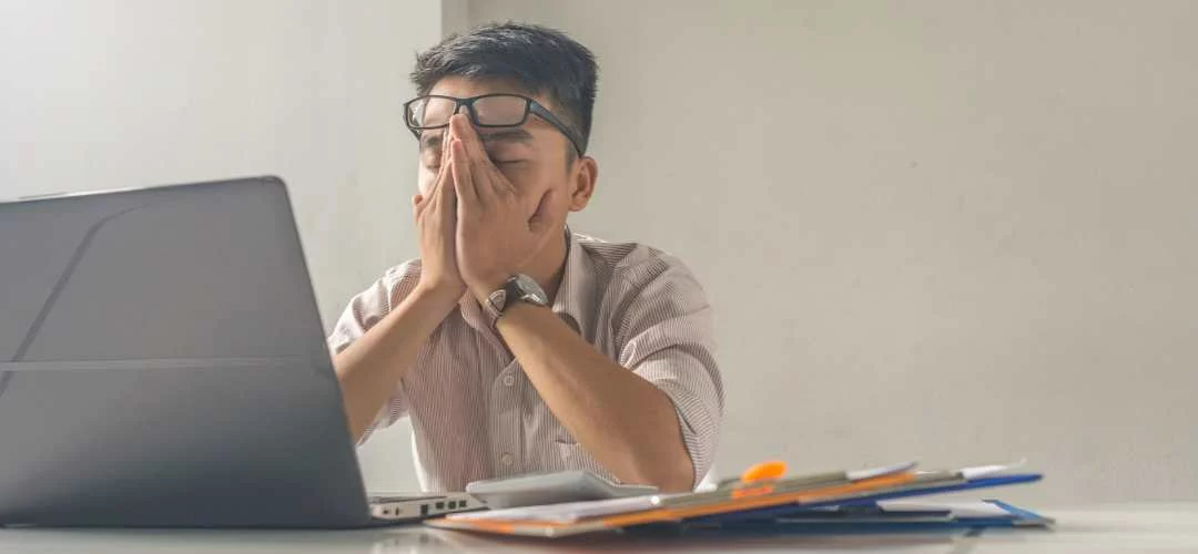 A man with glasses covering his face while working on a laptop.