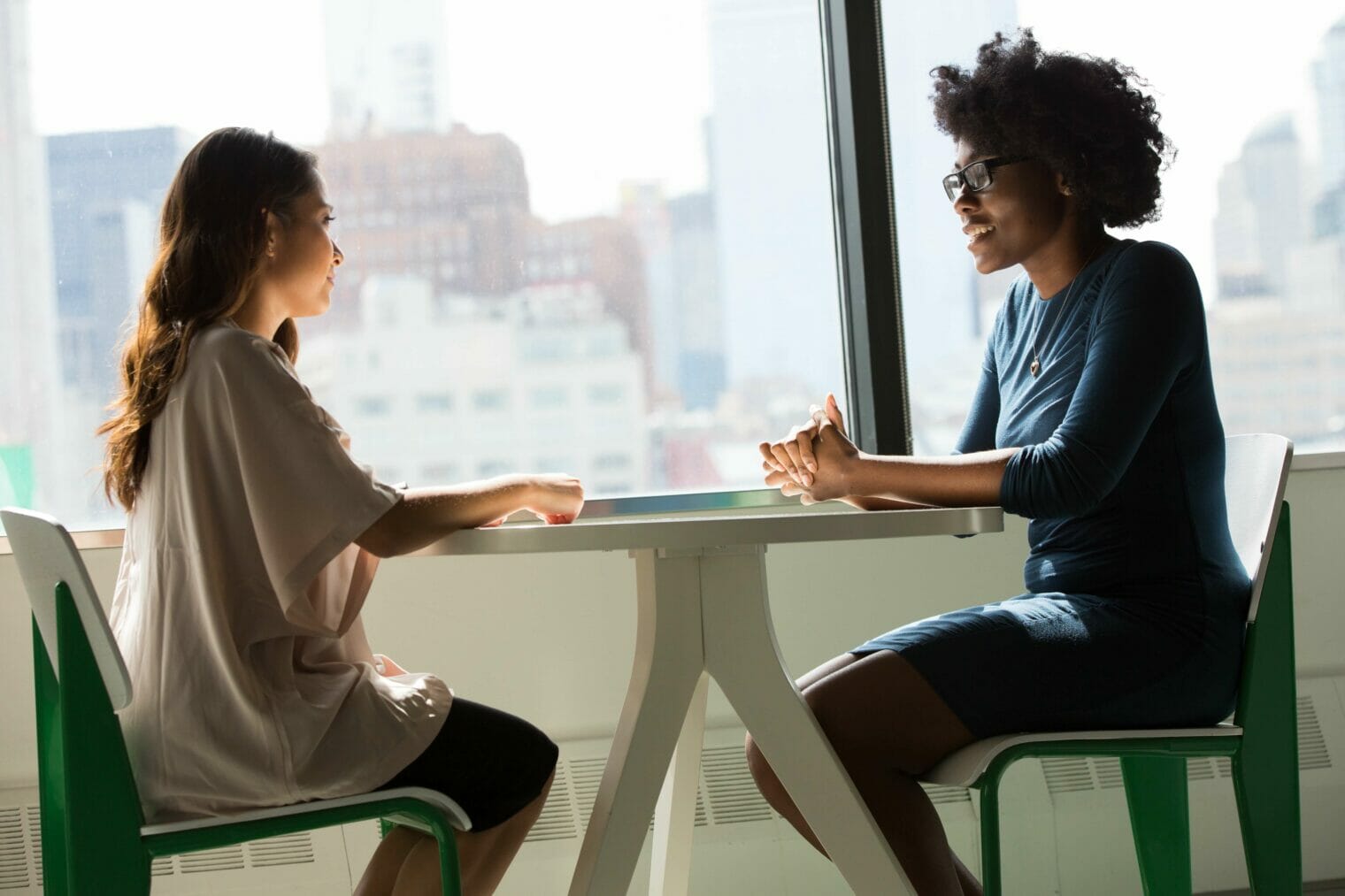 Two women engaged in confidential conversation at a table in an office.