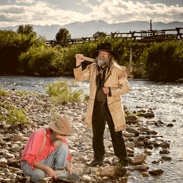 Two men dressed in traditional western attire by a rocky riverbank, one standing with a pickaxe, the other squatting with a pan.