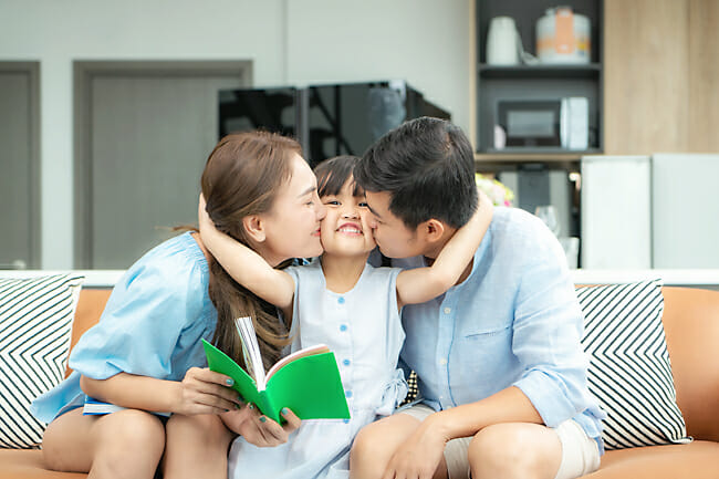 Parents kissing their happy child on the cheeks while sitting on a sofa, holding a book.