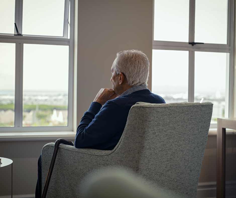 An elderly man sitting in a chair looking out a window, reflecting on his solitude.